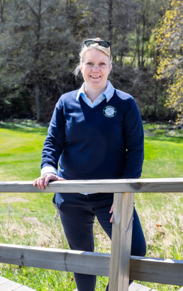 Bild på Lena Björkdahl som är klubbchef för Köpings Golfklubb. Hon står utomhus vid golfbanan.